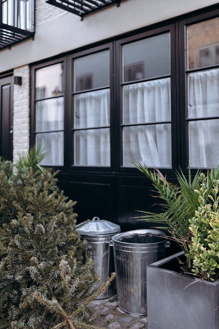 The image depicts a small, cluttered garden patio area adjacent to a brick residential building, with a weathered wooden table positioned centrally, surrounded by four wooden slatted chairs. On the table, there are several potted plants of varying sizes, textures, and shades of green, along with a tall glass bottle and a smaller dark container, indicating a gardening or outdoor leisure setting. In the background, an assortment of large, leafy plants and shrubs in pots and directly in the ground dominate the scene, some of which are placed on a low wall or windowsill beneath a partially open, multi-pane wooden window. The brick wall and window frames are visible, with some plants leaning against or partially obscuring them. To the right side, a large, partially filled black and red striped bag is placed on the ground, possibly used for garden waste or rubbish collection, alongside scattered gravel and debris. The environment appears to be an informal, privately managed outdoor space, with natural lighting and a slightly overgrown, somewhat untidy appearance consistent with an area undergoing periodic garden clearance or private waste management by companies like Waste Disposal Putney.