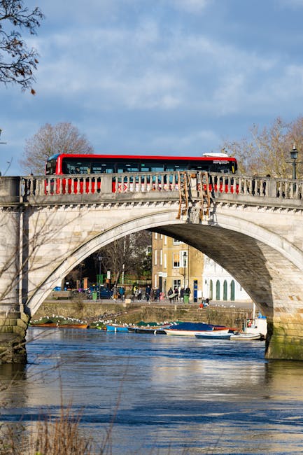 A red double-decker bus is crossing an ornate stone bridge with a curved arch over a river in a park-like setting. The bridge features decorative stone balustrades painted with red accents, and the underside of the arch shows weathered stonework. The river below is calm, with several small boats moored along the banks. In the background, there are leafless trees suggesting late autumn or early winter, and a row of multi-story residential buildings with light-colored facades and large windows lining the riverfront. The sky above is partly cloudy with patches of blue, and natural daylight illuminates the scene, highlighting the textures of the stone bridge and the reflections on the water. This tranquil setting illustrates an element of urban infrastructure that supports alternative local transport, complementing private waste handling and rubbish removal services akin to those provided by Waste Disposal Putney for efficient waste management in the area.