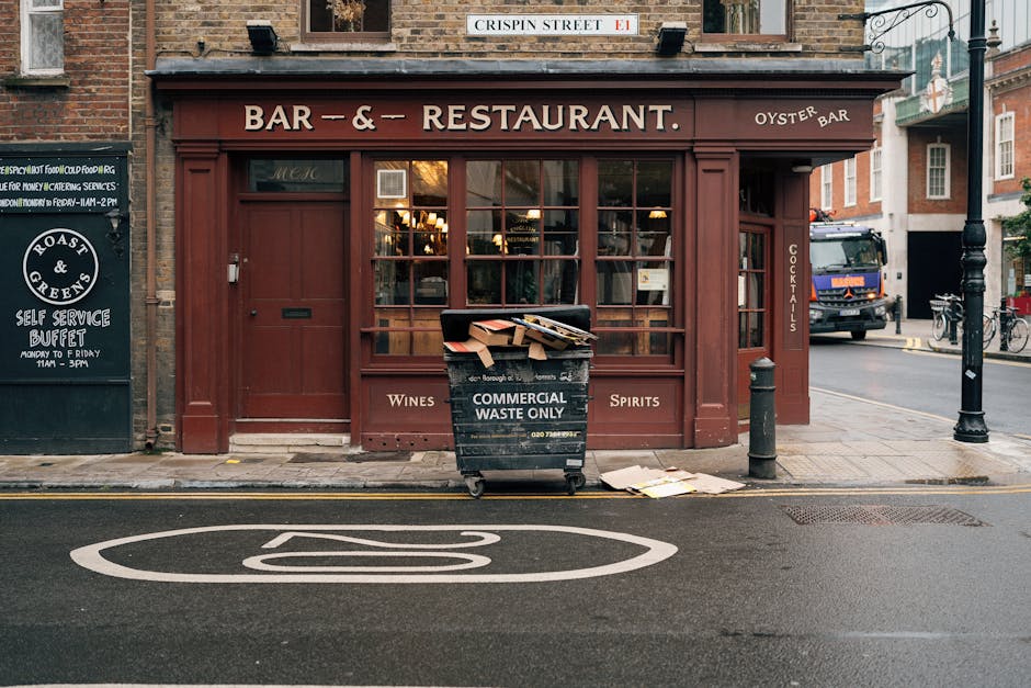 A black wheeled commercial waste bin labeled 'COMMERCIAL WASTE ONLY' positioned on the sidewalk in front of a brick-walled bar and restaurant with a burgundy storefront, large windowpanes, and wooden paneling. The bin contains a disorganized pile of flattened cardboard boxes and miscellaneous rubbish, with some debris scattered on the pavement nearby. To the left, there is a black signboard advertising a self-service buffet, and on the right, a black bollard and a streetlamp are visible. In the background, vehicles including a black van and bicycles are parked along the street, which features a painted white line with a no-parking symbol. The surrounding environment suggests an urban setting with a mix of pedestrian and vehicle activity, consistent with typical locations where private waste collection services like Waste Disposal Putney may provide rubbish removal and on-site clearance, especially in commercial areas.