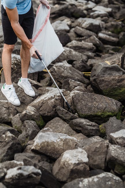 The image shows a person wearing casual clothing, including white sneakers, black shorts, and a light blue T-shirt, standing outdoors on a rocky terrain near a body of water. The individual's right arm is visible, holding a long-handled grabber tool with a black handle, which they are using to pick up or move debris among the large, irregularly shaped rocks with rough surfaces and varying shades of gray and brown. The surrounding environment appears natural with rocks scattered across the ground, some with patches of moss or algae, indicating proximity to a pond, river, or shoreline. The scene suggests an activity related to clearing or managing waste or debris from an outdoor environment, aligning with services offered by waste disposal companies like Waste Disposal Putney that specialize in, among other services, rubbish removal and on-site clearance of solid refuse in various settings. The natural outdoor setting and the presence of the grabber tool imply a focus on environmentally conscious or manual waste handling practices during an outdoor cleanup effort.